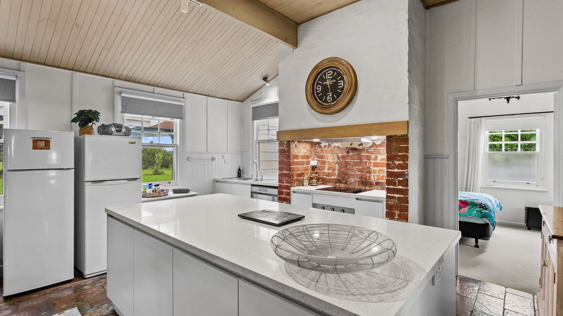 Una cocina moderna con armarios blancos, una pared de acento de ladrillo, una isla grande con un bol de vidrio y un reloj en la pared.