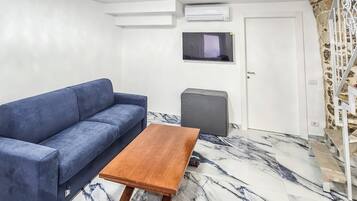 Une pièce avec un canapé bleu, une table en bois, un téléviseur et un escalier adossé à un mur en pierre.