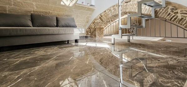 Una sala moderna con piso de mármol, un sofá gris y una pared de piedra.