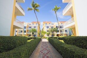A hotel with a central pathway flanked by neatly trimmed hedges and palm trees, leading to a multi-story building with balconies. - Estrella Del Mar F4_400Mbps_BBQ_Pool_Walk to Beach (Punta Cana)