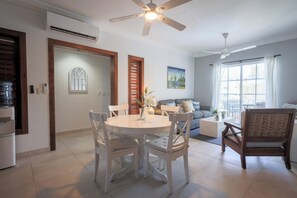 A modern kitchen with white cabinetry, a dining area with a round table and white chairs, a living room with a sofa and a painting, and a ceiling fan. - Estrella Del Mar F4_400Mbps_BBQ_Pool_Walk to Beach (Punta Cana)