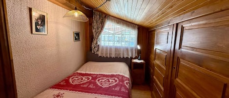 A bedroom with a wooden ceiling, a bed with a red and white bedspread, a nightstand with a lamp, and a window with lace curtains.