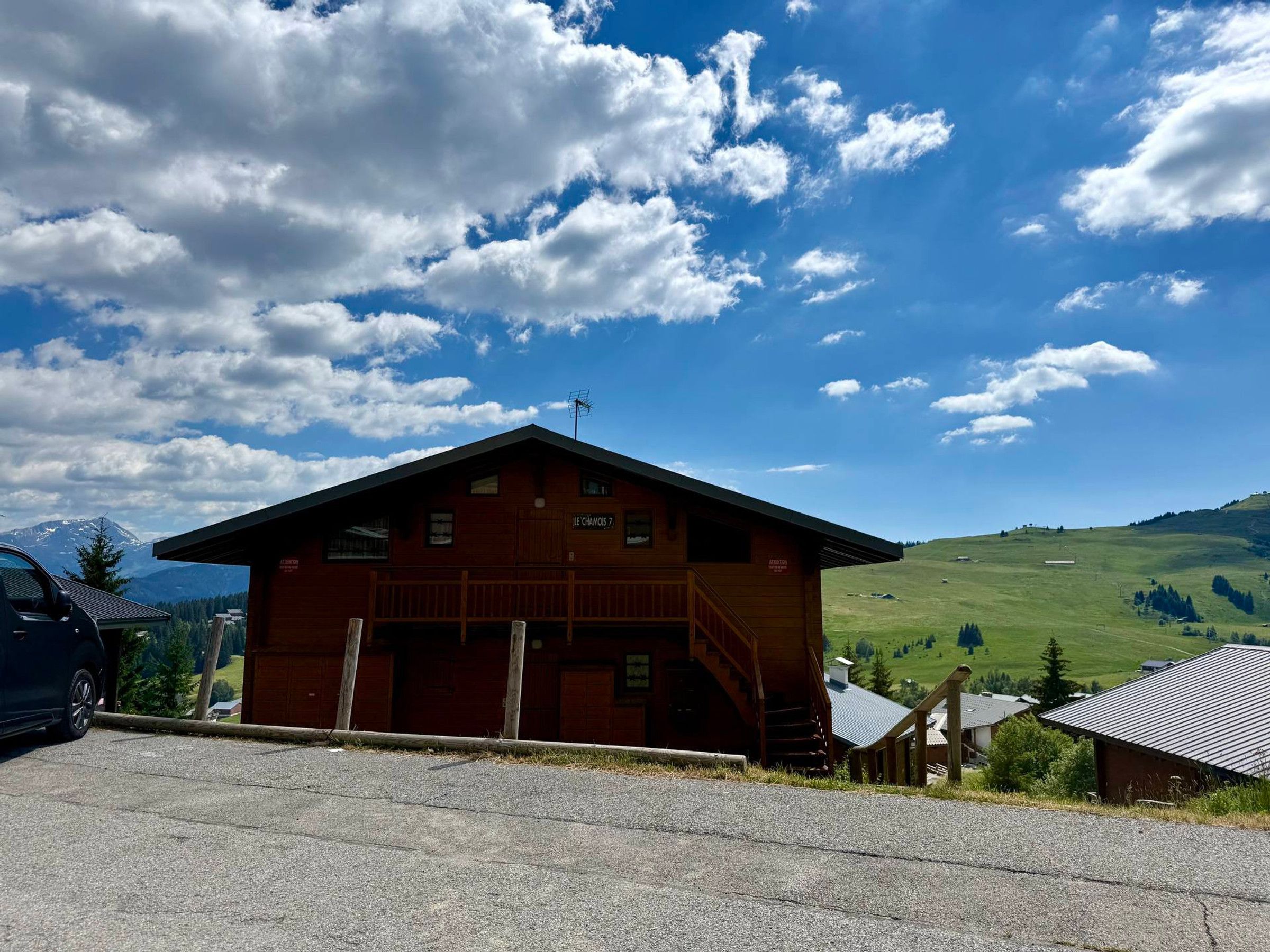 Un bâtiment en bois doté d’un toit en pente, d’un escalier menant à l’entrée et offrant une vue sur une vallée verdoyante, avec des montagnes en arrière-plan.