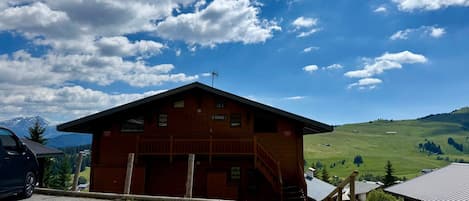 A wooden building with a sloped roof, a staircase leading to the entrance, and a view of a green valley with mountains in the background.