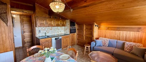 A cozy cabin interior with a wooden ceiling, a dining table set for two, a sofa, and a kitchen area.