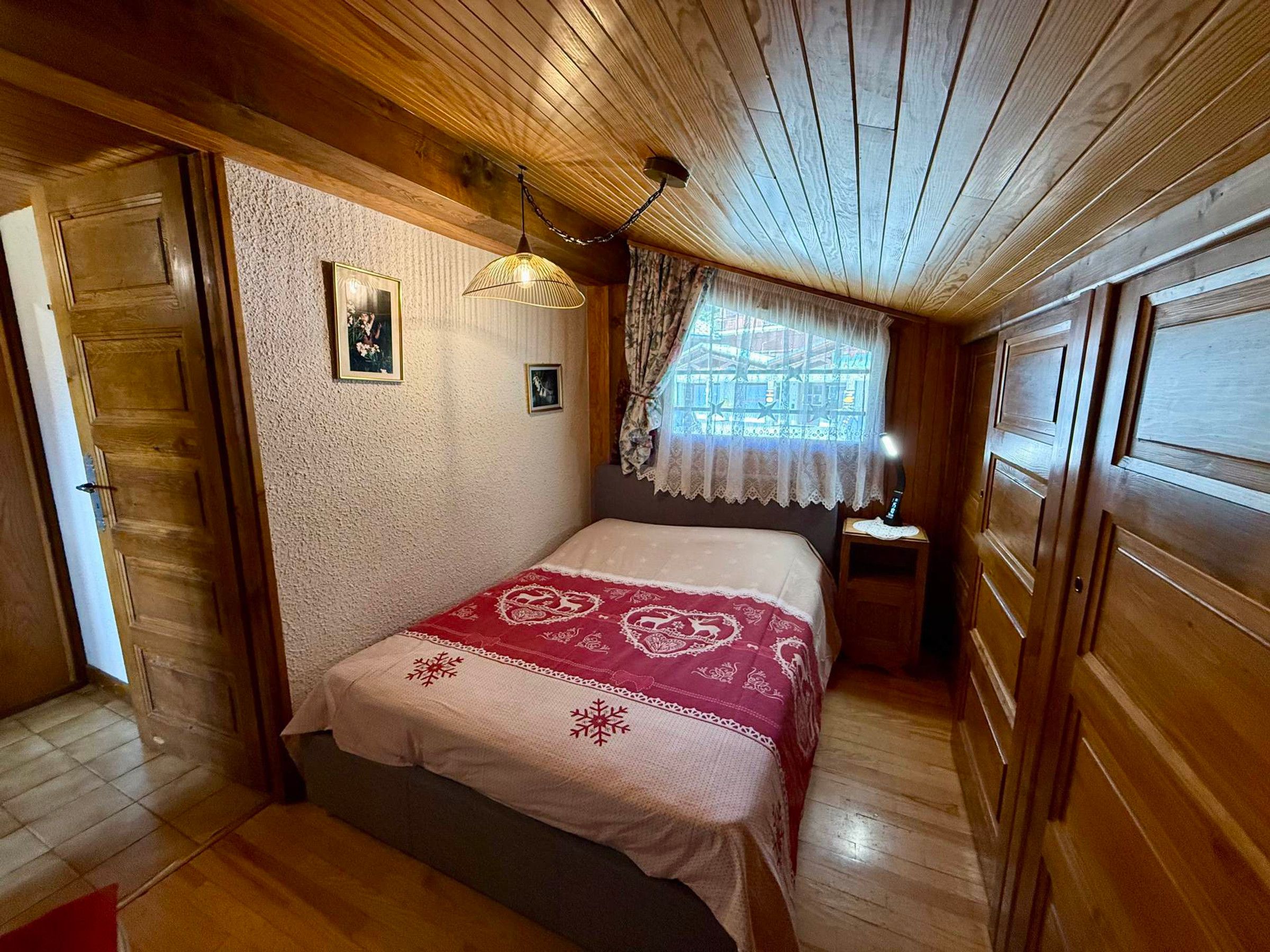 Een gezellig slaapkamer met een houten plafond, een bed met een beddegoed met dessin, een nachtkastje met een lamp en een raam met een kantelaar.