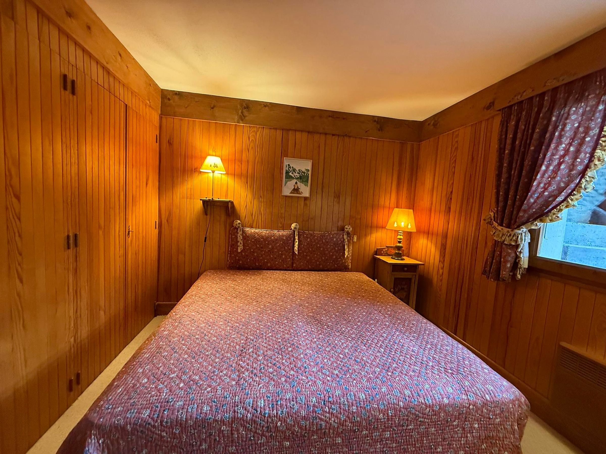 A wooden-paneled room with a bed, two bedside lamps, and a window with curtains.