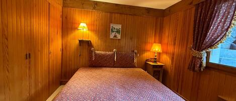 A wooden-paneled room with a bed, two bedside lamps, and a window with curtains.