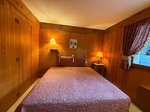 Ein Zimmer mit Holzwandverkleidung, einem Bett, zwei Nachttischlampen und einem Fenster mit Vorhängen.