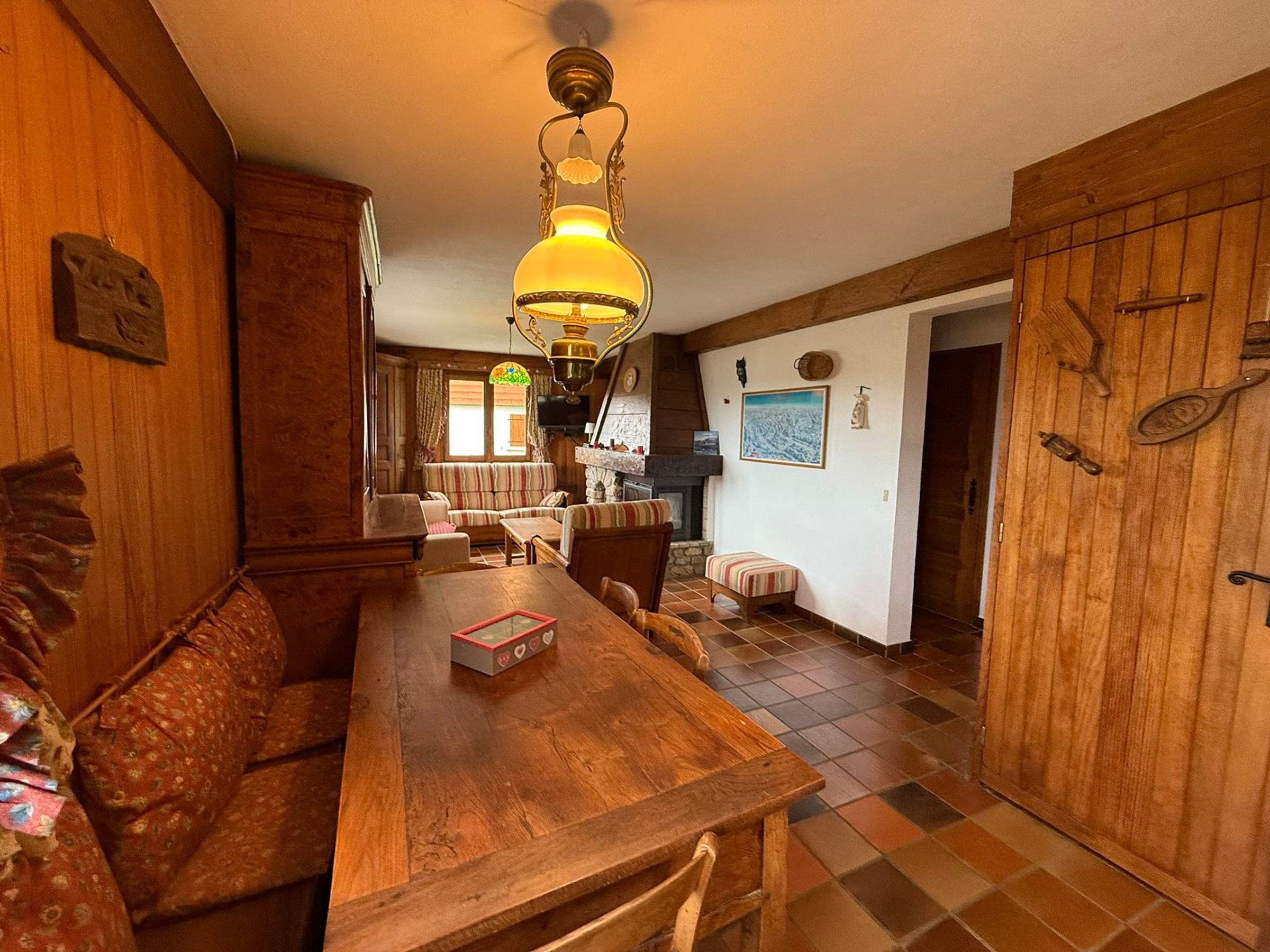 A cozy wooden interior with a dining table, chairs, and a bench. A fireplace is visible in the background, and there are wooden cabinets and a tiled floor.