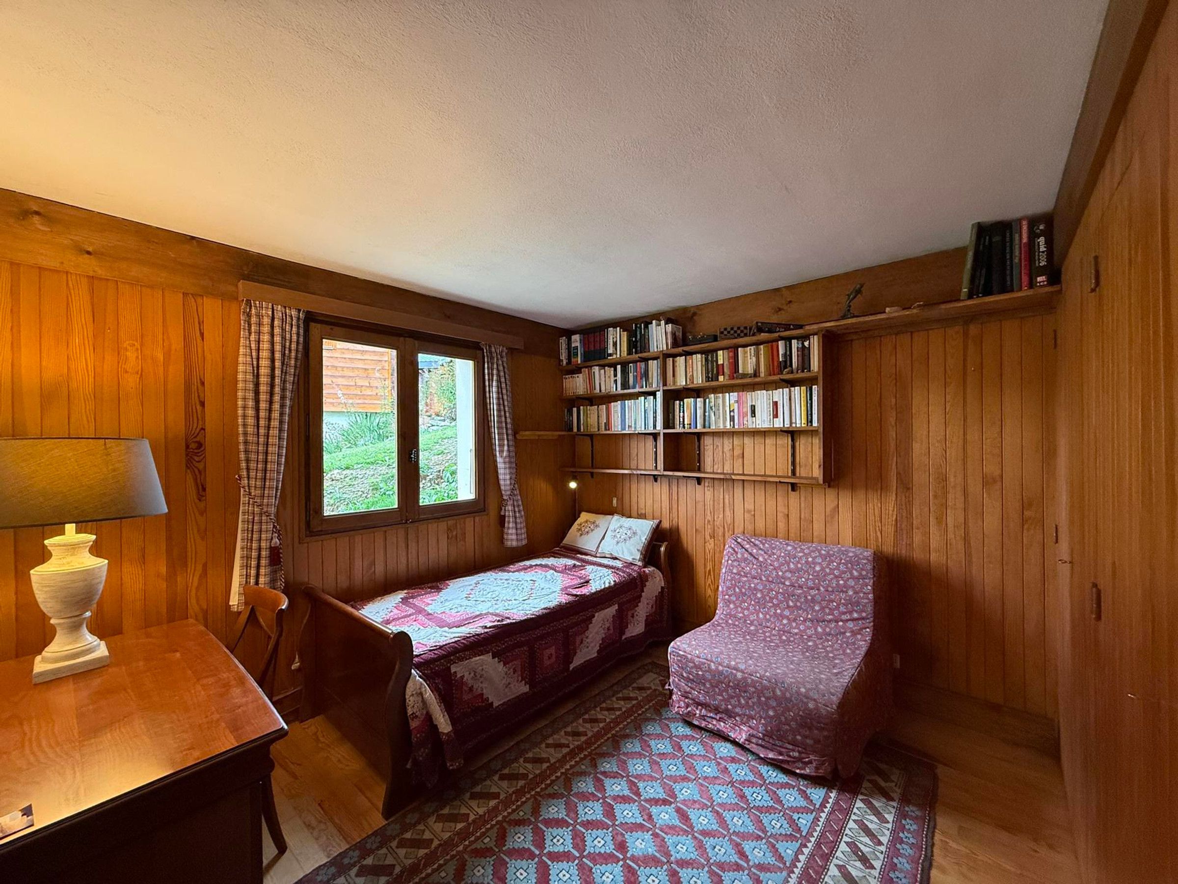 A cozy room with a bed, a desk, a chair, a bookshelf, and a window with curtains.