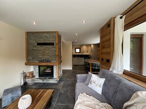 Ein modernes Wohnzimmer mit Kamin aus Stein, einer Couch, einem Couchtisch und Blick in die Küche im Hintergrund.