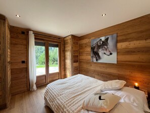A bedroom with wooden walls, a large window, a bed with white bedding, and a framed picture of a wolf. - Cozy apartment, mountain view - 6 guests (La Clusaz)