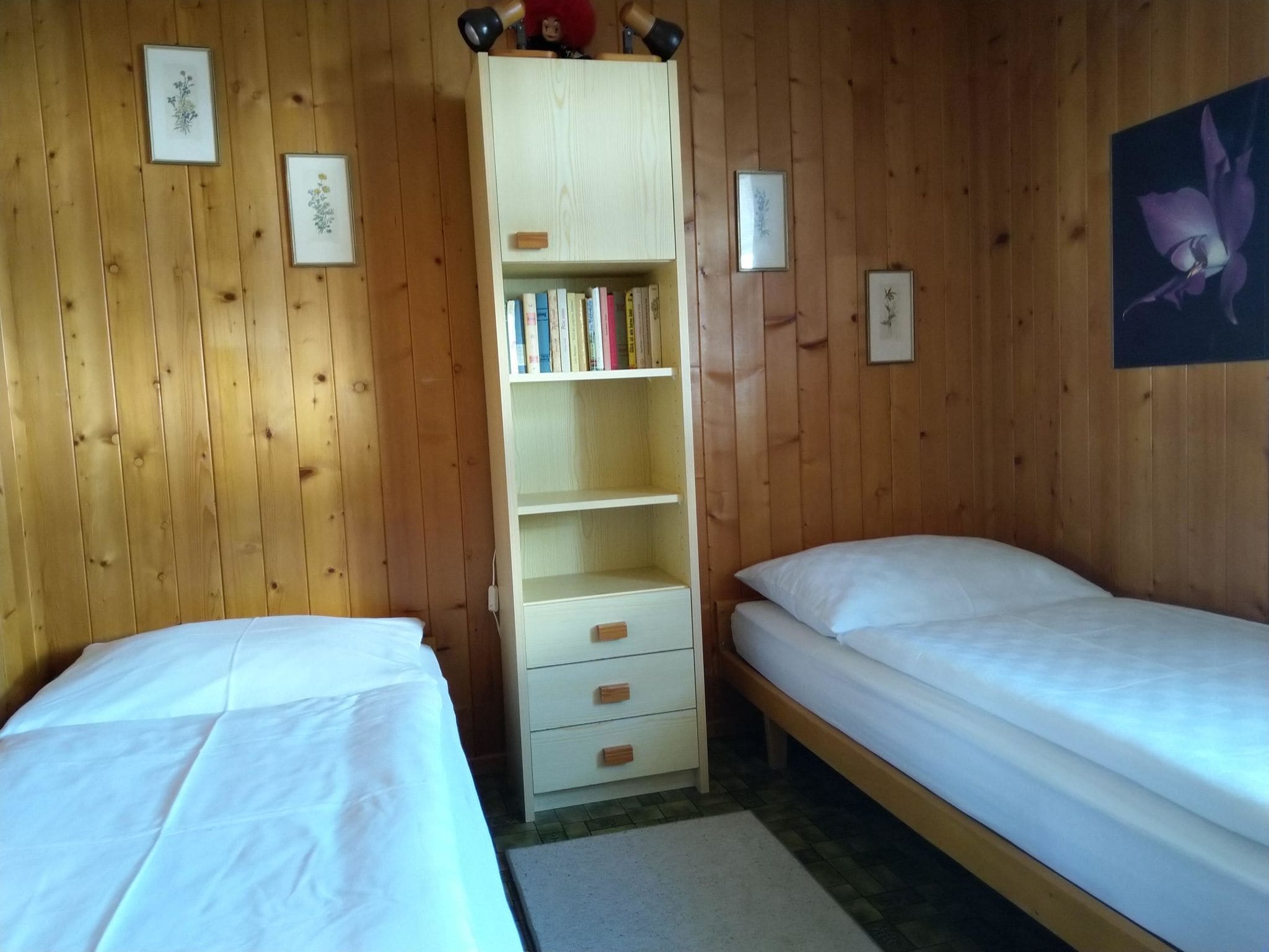 A room with two beds, a wooden wardrobe, and framed pictures on the walls.