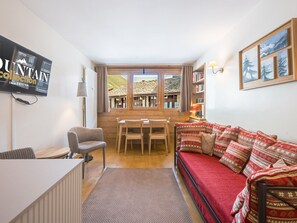 A cozy living room with a red sofa, a wooden dining table with chairs, a bookshelf, and a large window with a view of a mountain. - Apartment at the slopes - 4 guests (Val-d'Isère)