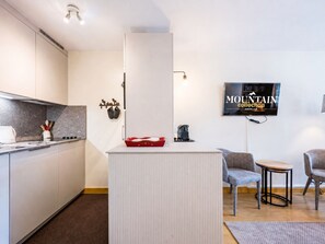A modern kitchen with a built-in stove, a coffee machine, and a red tray on the counter. There is a seating area with two chairs and a small table. A television is mounted on the wall displaying 'MOUNTAIN Collection'. - Apartment at the slopes - 4 guests (Val-d'Isère)