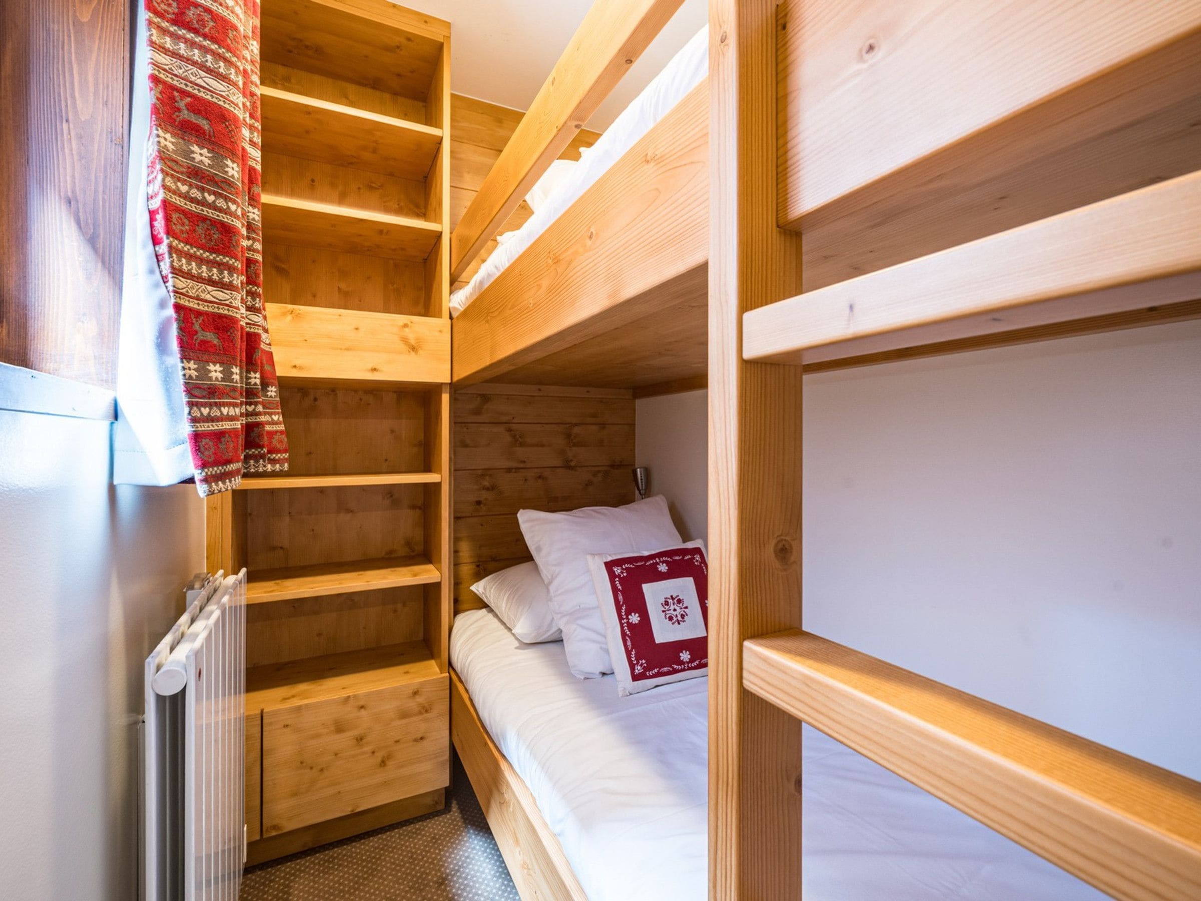 Quarto pequeno com beliche, prateleiras de madeira e radiador.