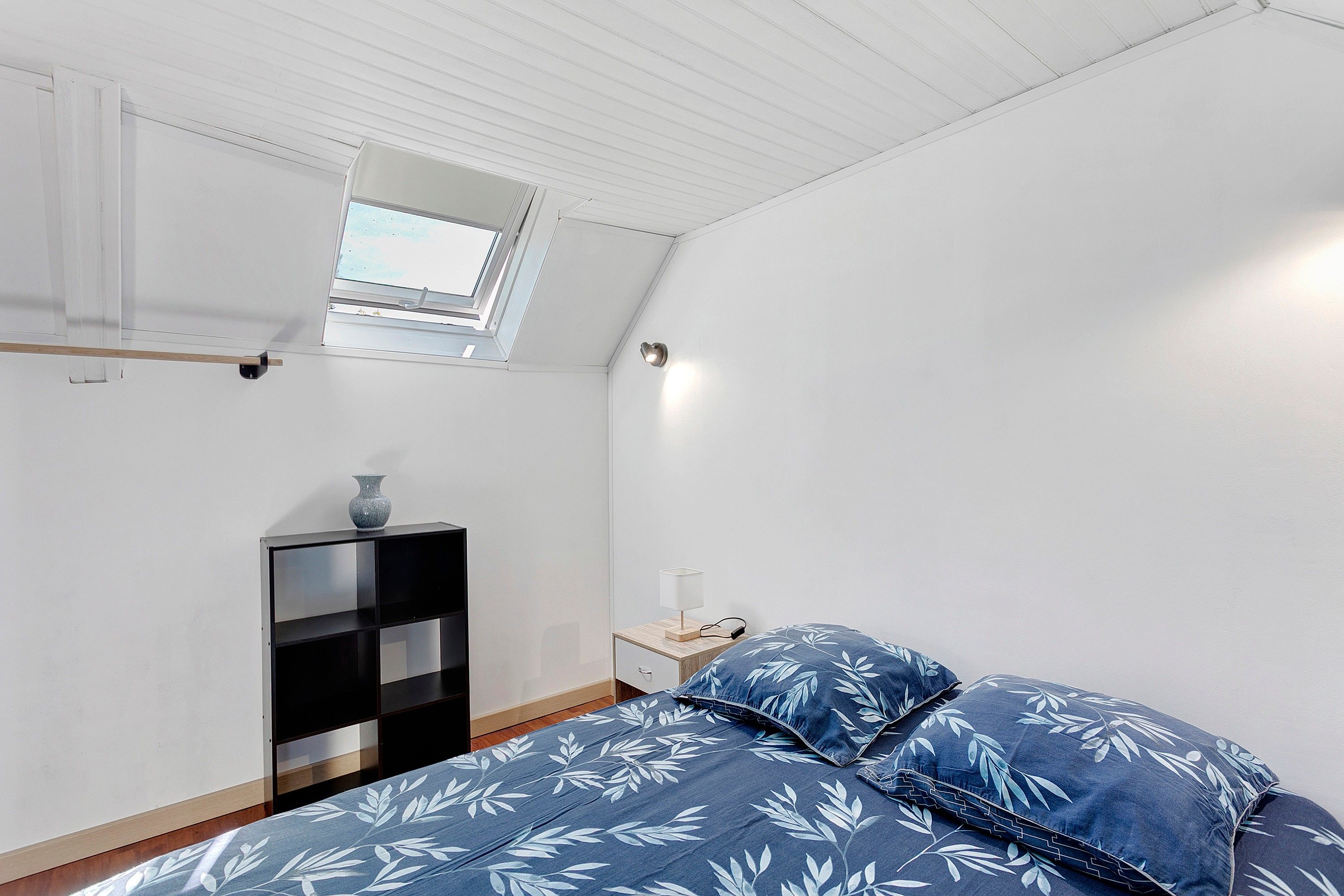 A bedroom with a skylight, a bed with blue floral bedding, a bedside table with a lamp, and a black storage unit.
