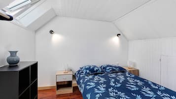 Ett sovrum med ett sluttande tak, en säng med ett täcke med ett lövmönster, sängbord med lampor, ett litet träskåp och en vas på en hylla.