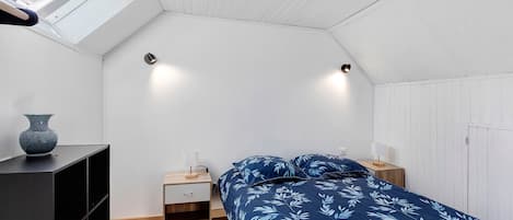 Een slaapkamer met een schuine kap, een bed met een beddegoed met bladmotief, nachtkastjes met lampen, een klein houten kastje en een vaas op een plank.