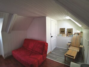 Ett rum med en röd soffa, ett litet bord och en bänk. På en hylla står böcker och inramade bilder.