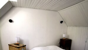 Een kleine eenpersoonskamer met schuine balken, een houten vloer, een wit bed, een vintage koffer en een klein houten nachtkastje met een lamp.