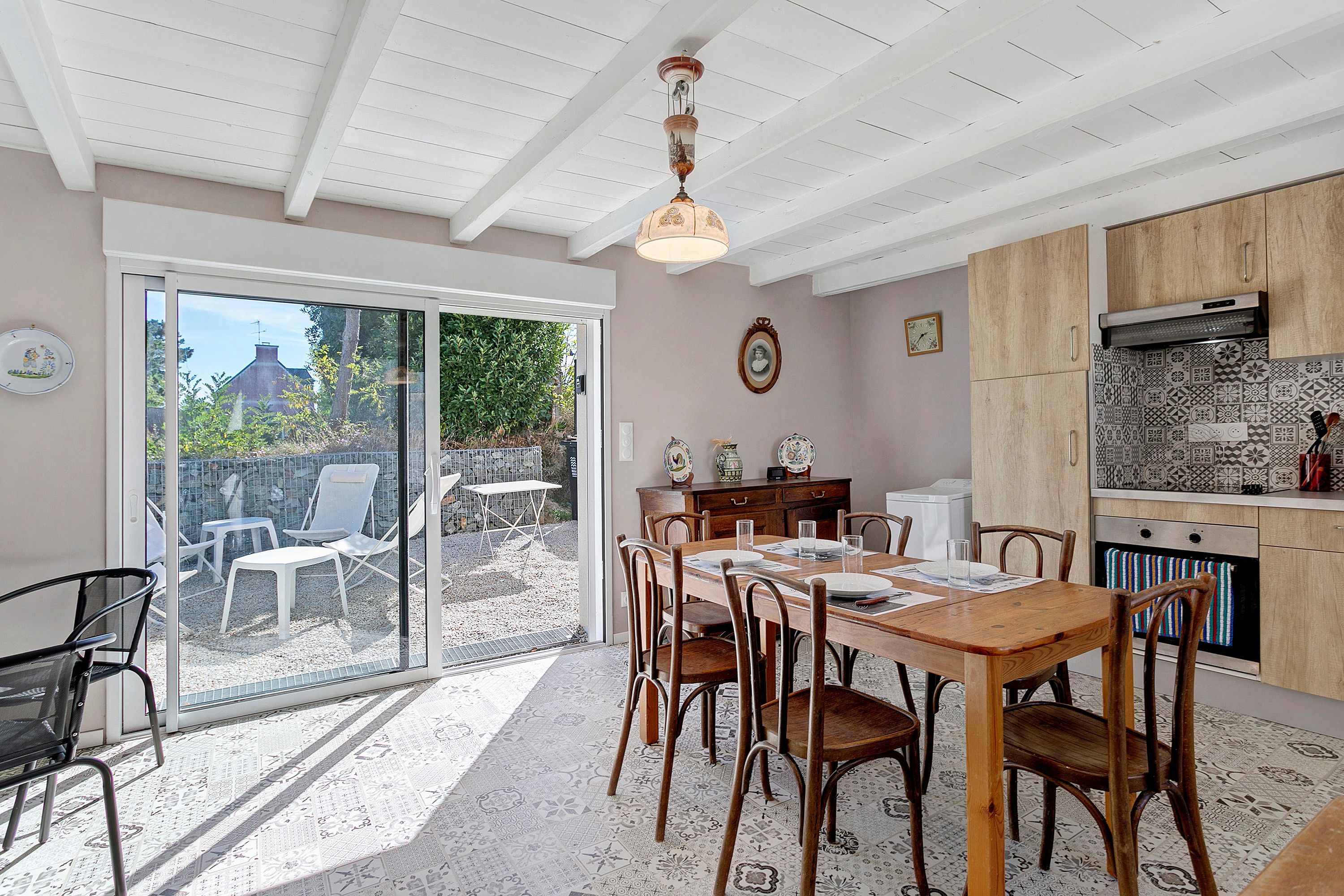 Un coin repas avec une table dressée pour quatre personnes, entourée de chaises, une cuisine avec des meubles en bois et une porte-fenêtre coulissante donnant sur un patio extérieur.