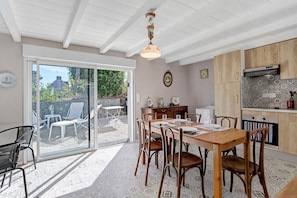 Uma sala de jantar com uma mesa posta para quatro pessoas, rodeada por cadeiras, uma cozinha com armários de madeira e uma porta de vidro deslizante que dá para um pátio externo.
