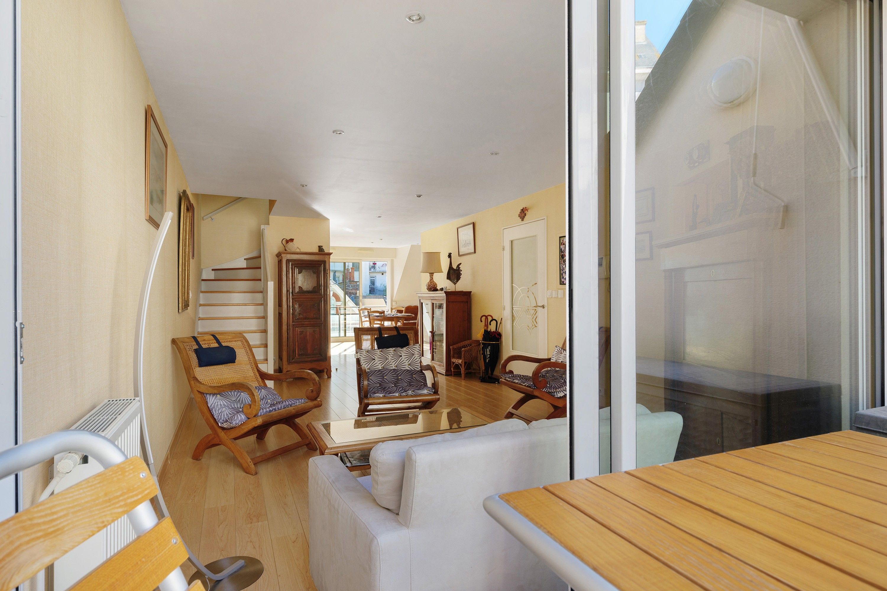 A spacious living area with a wooden table, two sofas, and a dining table. A staircase leads to an upper floor, and there is a door with a decorative handle.