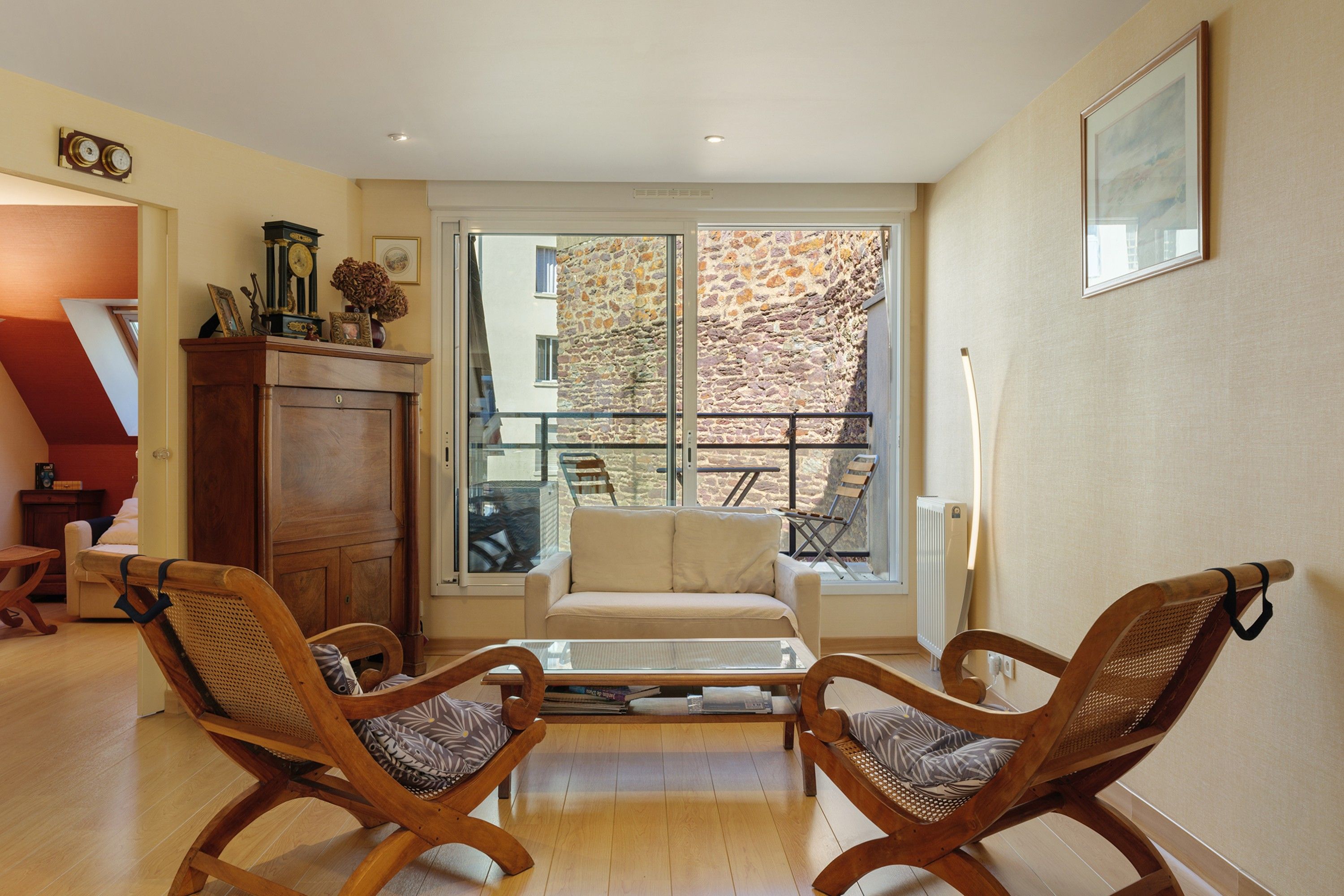 Ein Wohnzimmer mit einer Couch, zwei Holzstühlen, einem Couchtisch und einem großen Fenster mit Blick auf eine Steinmauer und ein Gebäude.