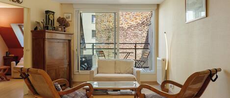 Ein Wohnzimmer mit einer Couch, zwei Holzstühlen, einem Couchtisch und einem großen Fenster mit Blick auf eine Steinmauer und ein Gebäude.