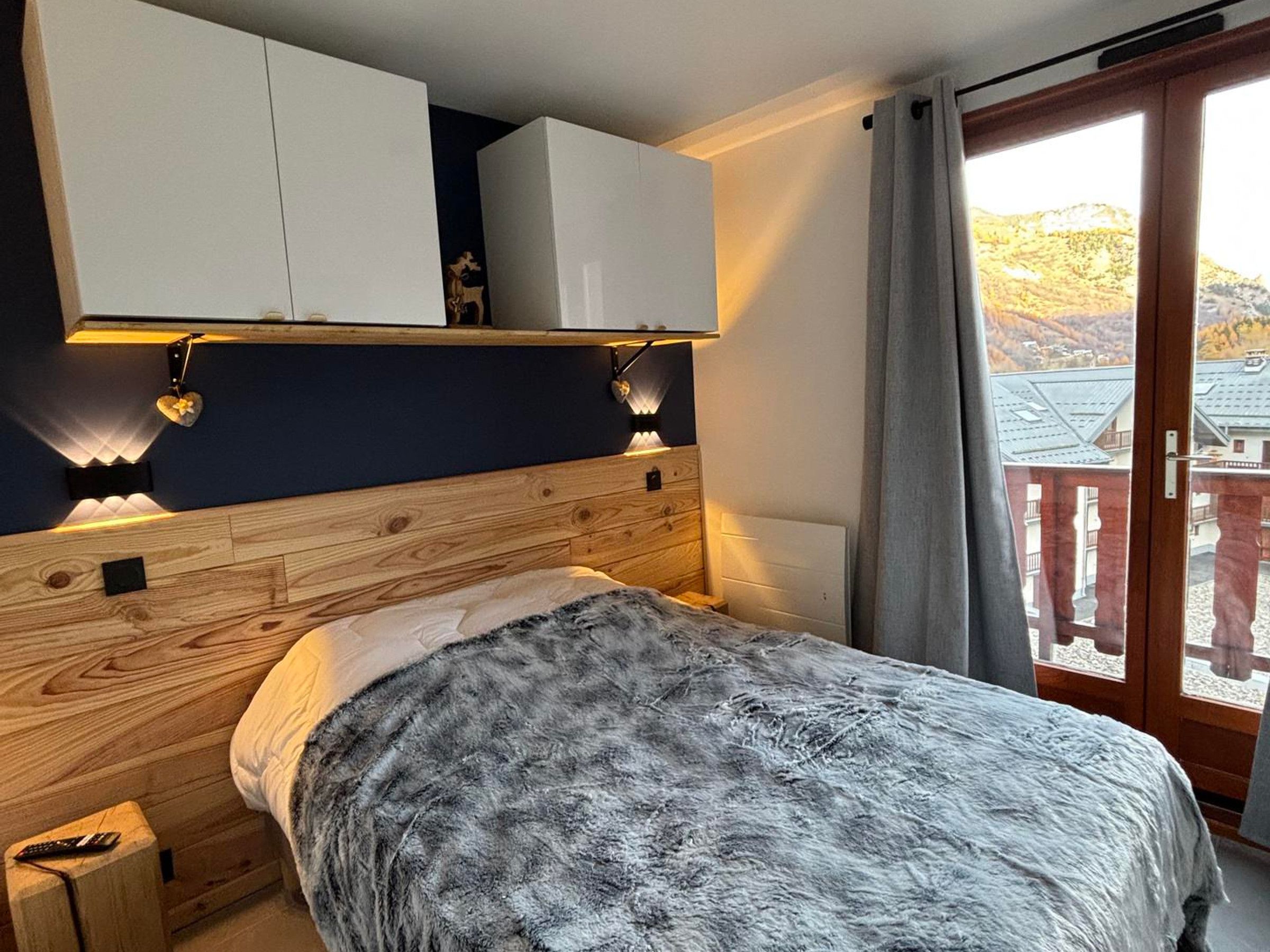 Un dormitorio con cama, cabecero de madera, armarios empotrados, luces fijadas a la pared y vistas a través de un ventanal.