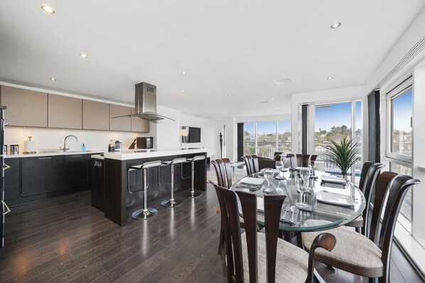 A modern kitchen with dark wood flooring, a central island with bar stools, and a dining area with a glass table and chairs. - The Residences Serviced Apartments (Aberdeen)