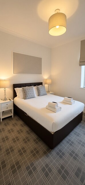 A hotel room with a large bed, two bedside tables, a wall-mounted lamp, and a window with blinds. - The Residences Serviced Apartments (Aberdeen)