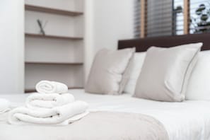 A neatly made bed with rolled white towels, a headboard, and a window with blinds.