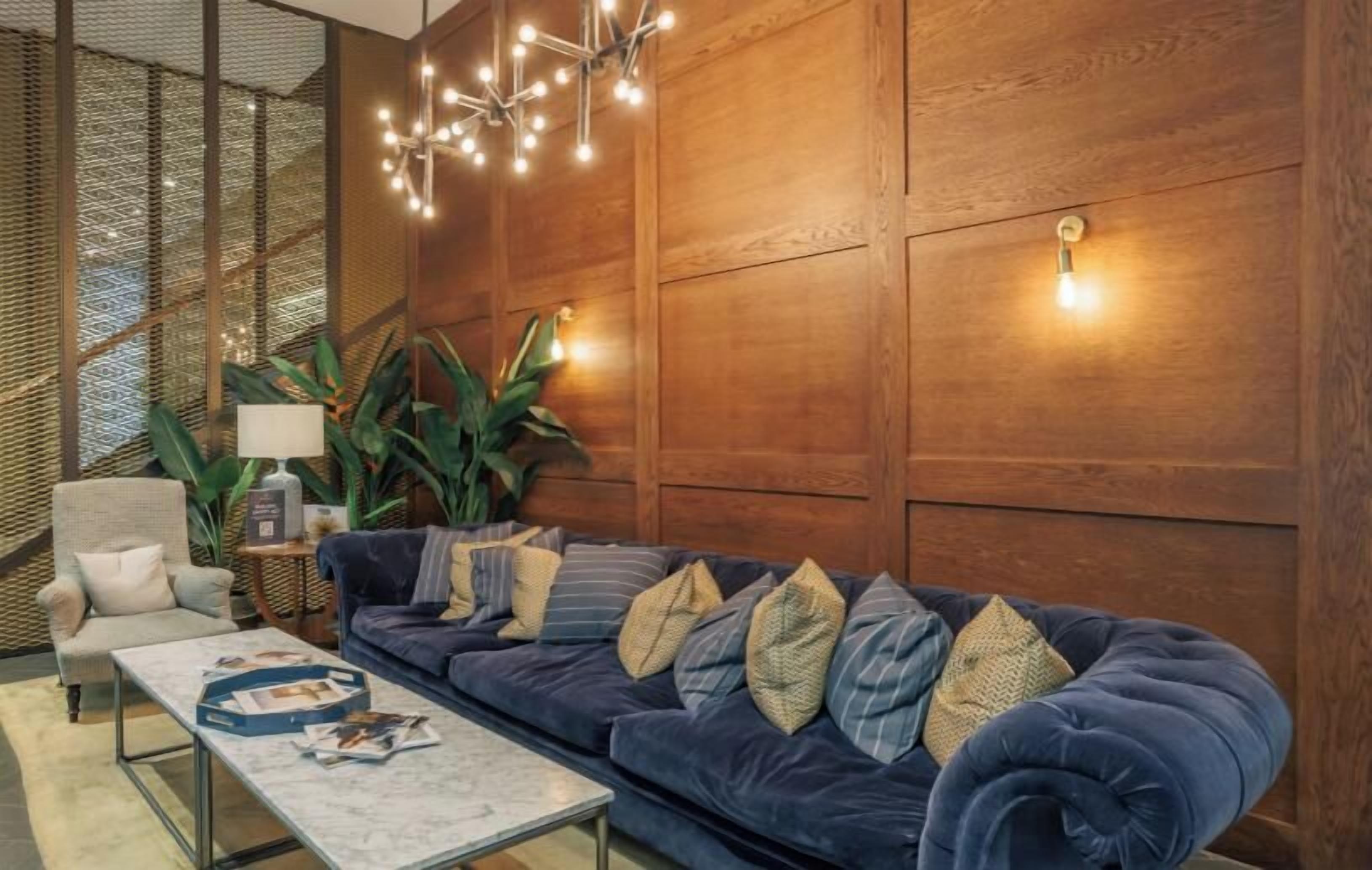 A modern living room with a dark blue sofa, a coffee table, and a lamp. There is a wooden wall paneling and a plant in the background.
