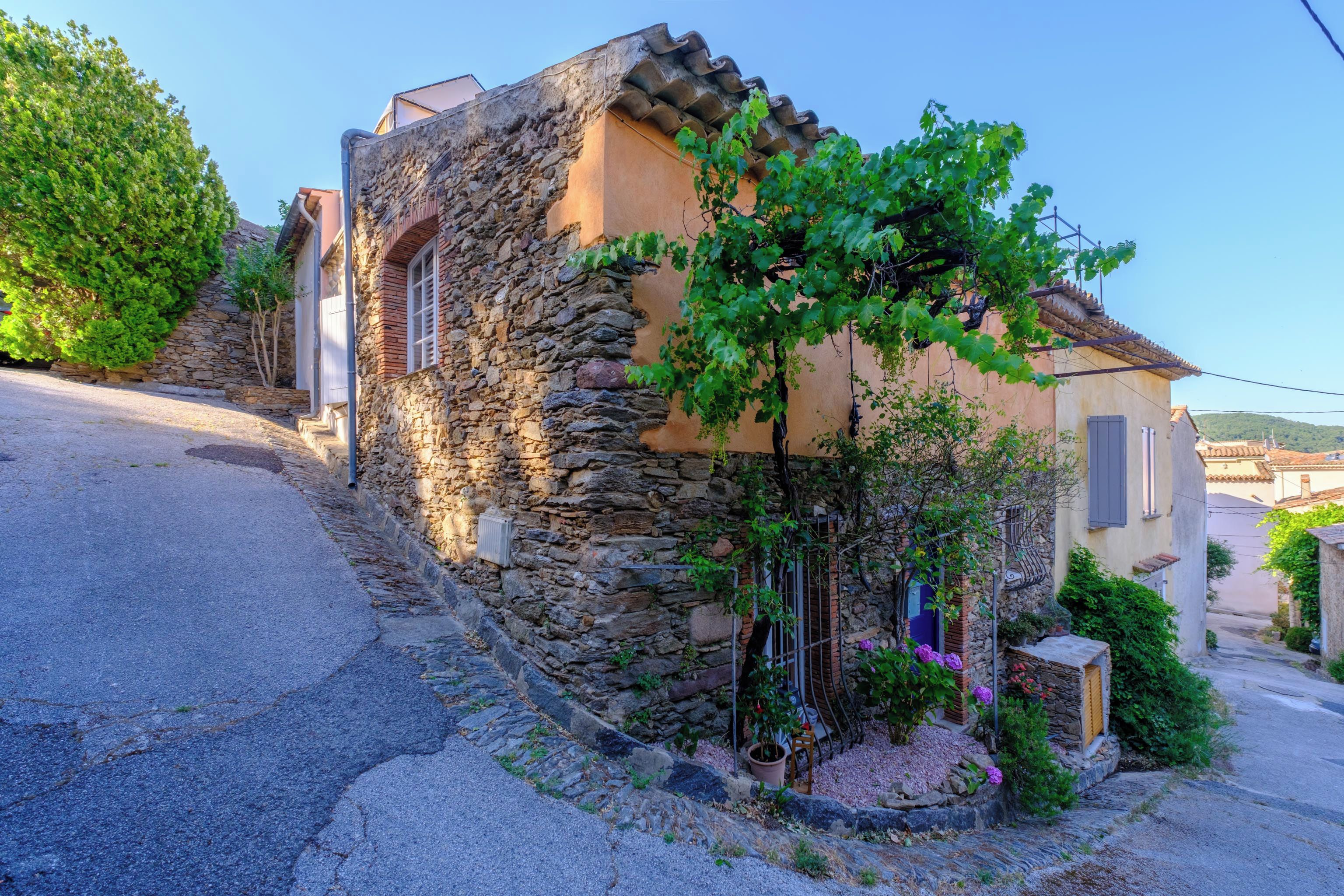 A stone building with a sloped roof, a small garden with a tree, and a paved street.