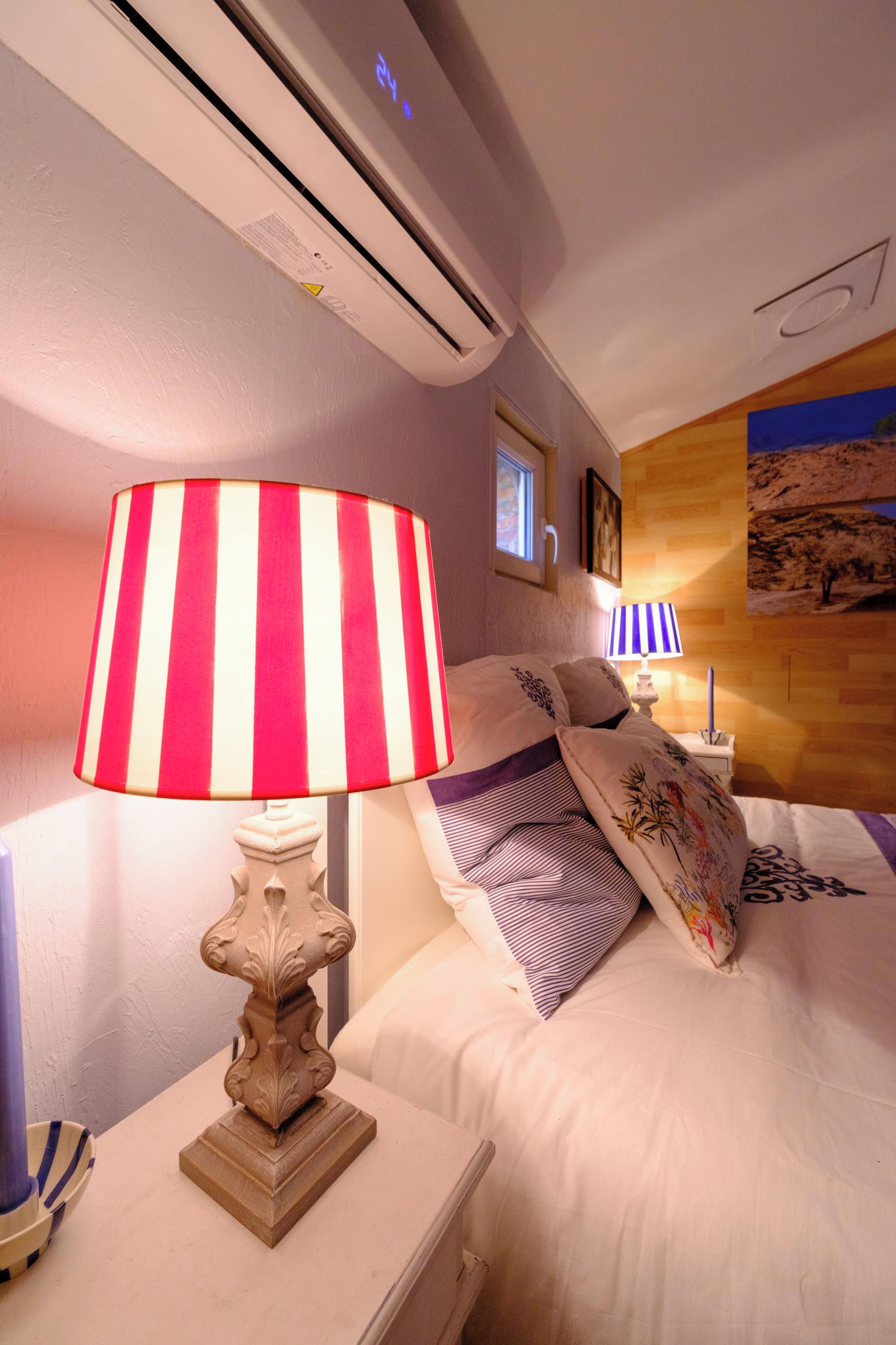 Habitación de hotel con cama, mesita de noche, lámpara, equipo de aire acondicionado, ventana y un cuadro colgado en la pared.