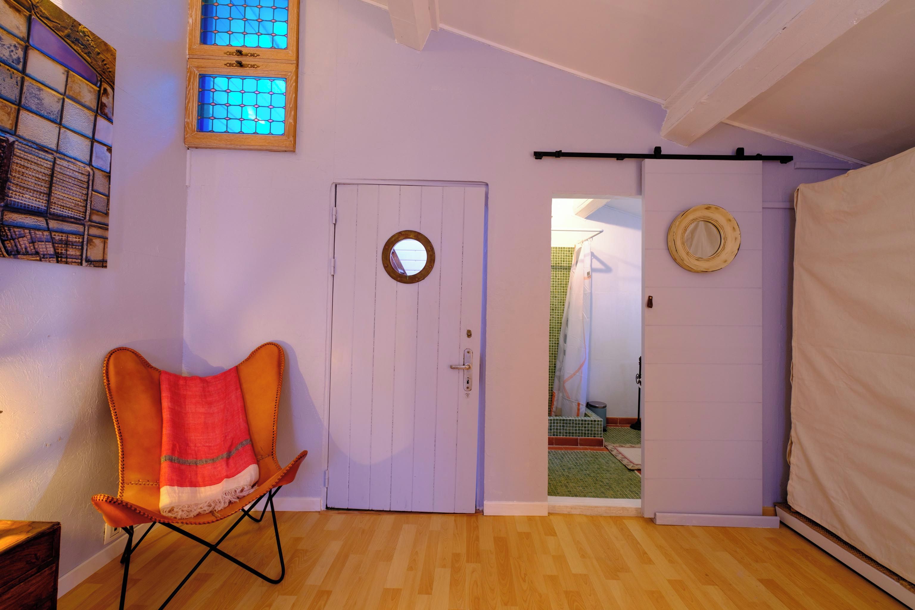 Een kamer met een houten vloer, een witte deur met een ronde spiegel, een klein raam met blauw getint glas, en een stoel met een rood-wit deken.