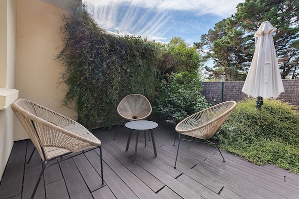 An outdoor seating area with wicker chairs and a table, surrounded by greenery and a white umbrella.