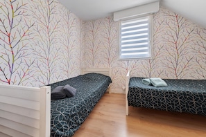 Un petit logement composé d’une seule pièce, comprenant deux lits simples, une fenêtre avec des stores et un mur recouvert d’un papier peint à motifs d’arbres.