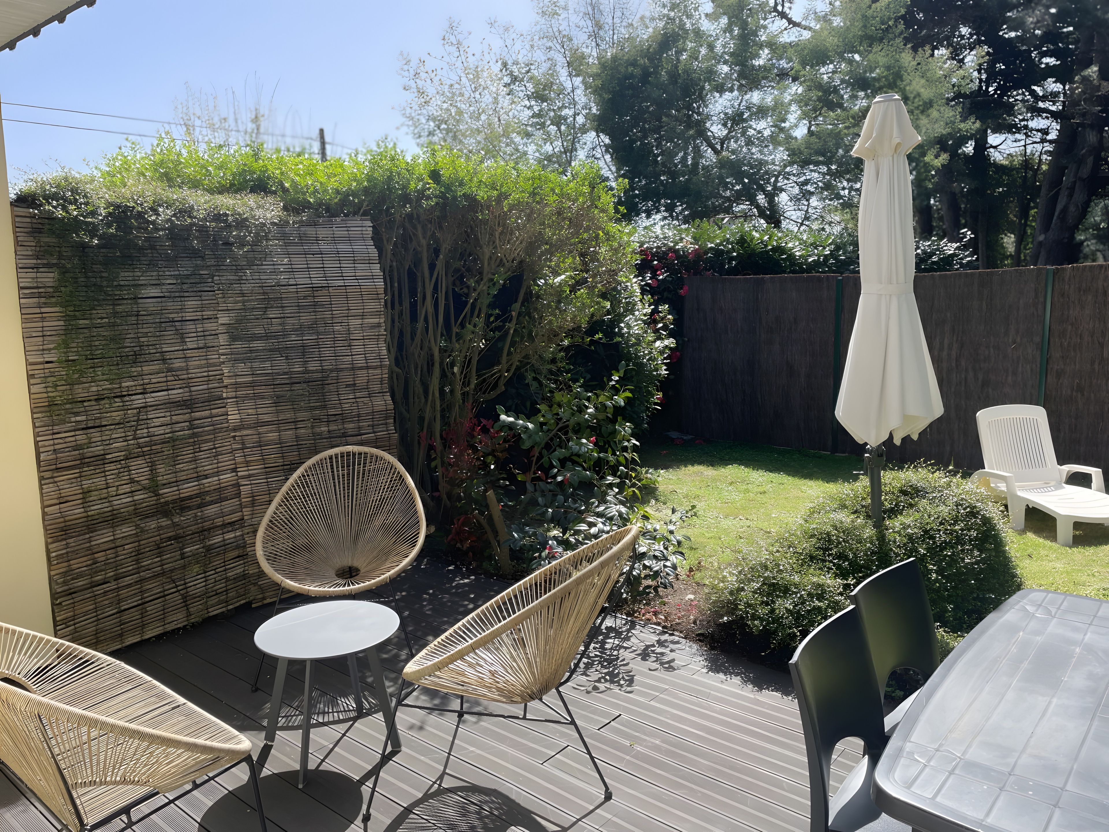 Eine Terrasse mit Korbstühlen und einem Tisch, umgeben von einer Holzdielenveranda und einem Gartenbereich mit Holzzaun und Grünpflanzen.
