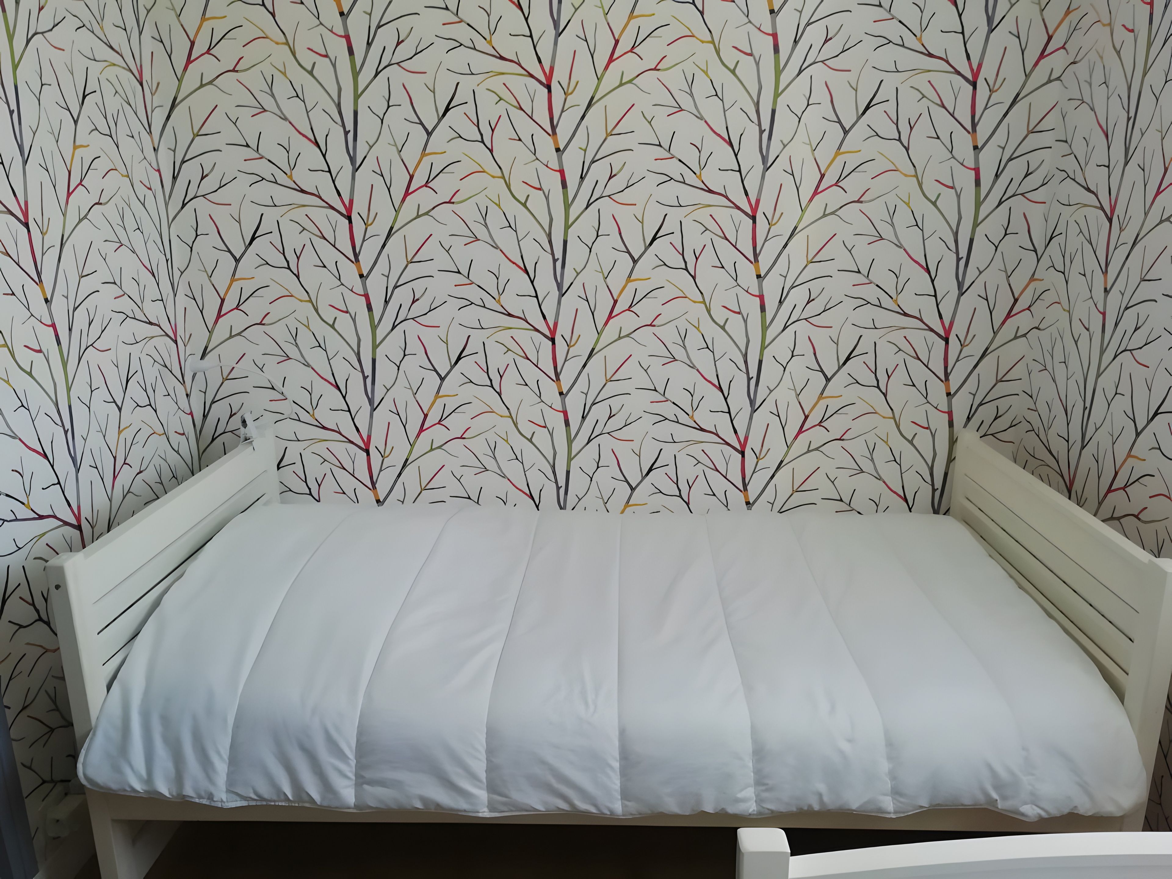 A bed with a white mattress and headboard against a wall with a tree branch pattern wallpaper.