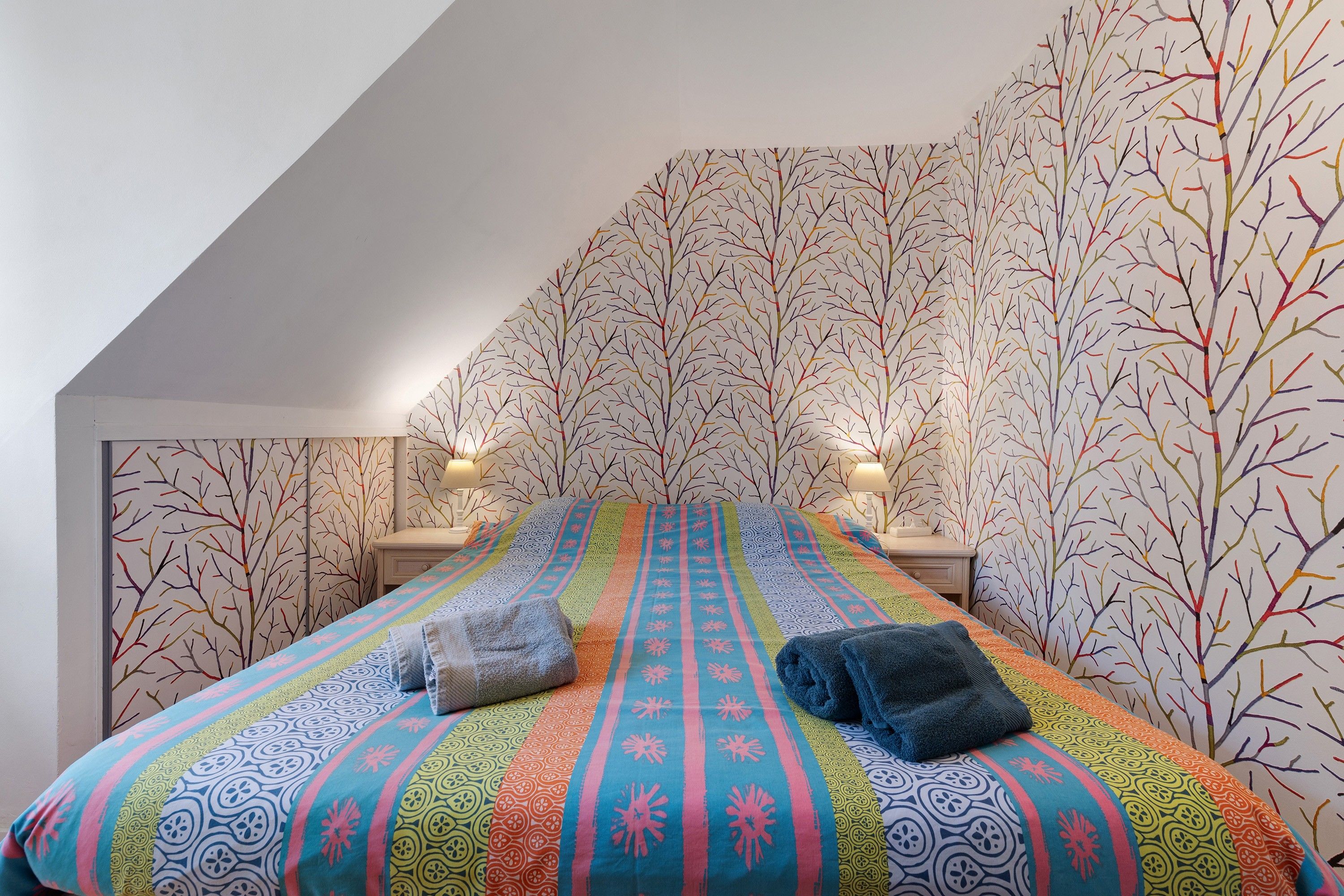 A bedroom with a sloped ceiling, a colorful bedspread, two bedside lamps, and a tree-patterned wallpaper.