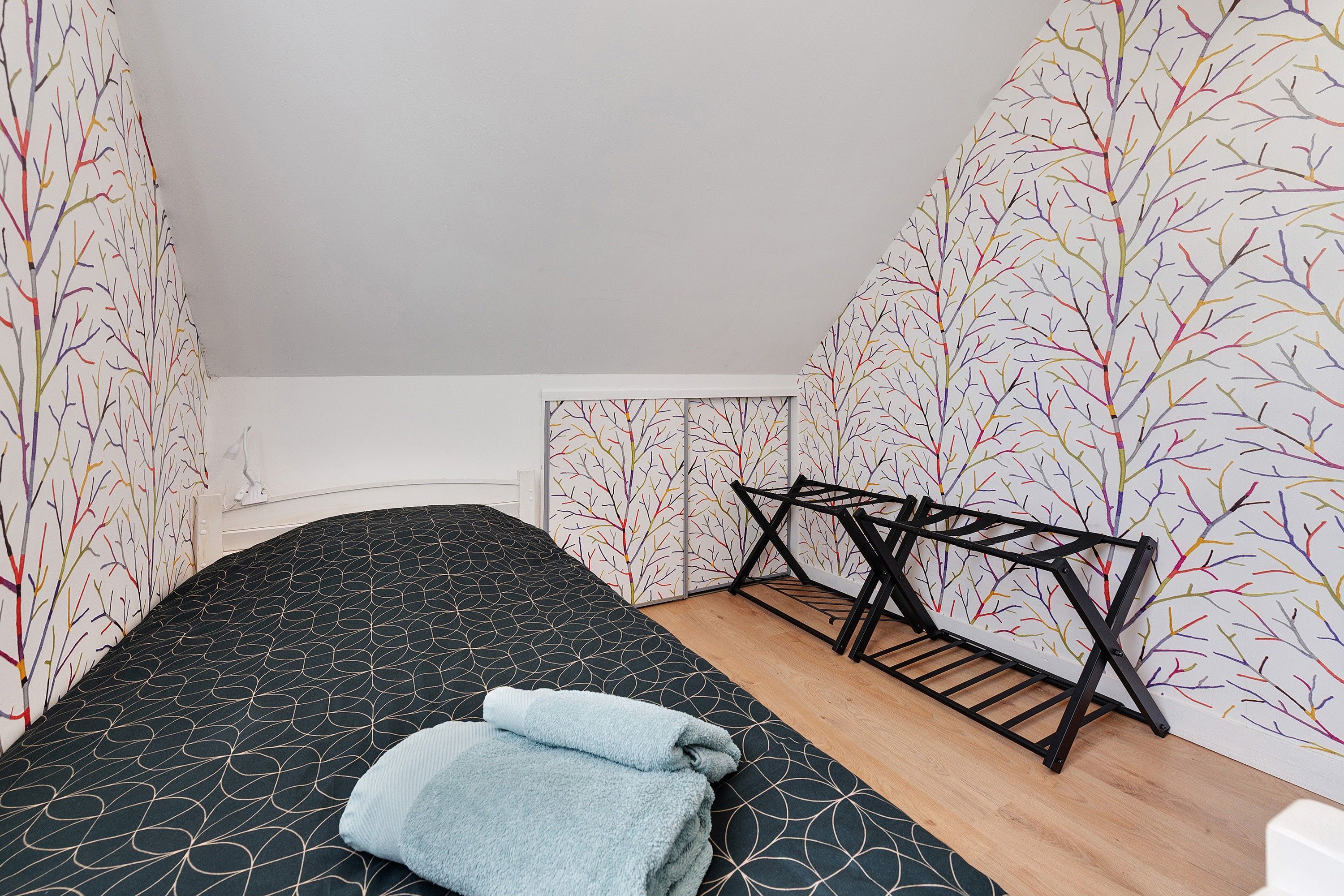 Een kleine kamer met een enkel bed, een zwart beddegoed met patroon, een houten vloer en een zwarte metalen bankje.