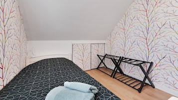 Een kleine kamer met een enkel bed, een zwart beddegoed met patroon, een houten vloer en een zwarte metalen bankje.