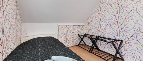 Ein kleines Zimmer mit einem Einzelbett, einer Bettdecke mit schwarzem Muster, Holzboden und einer schwarzen Metallbank.