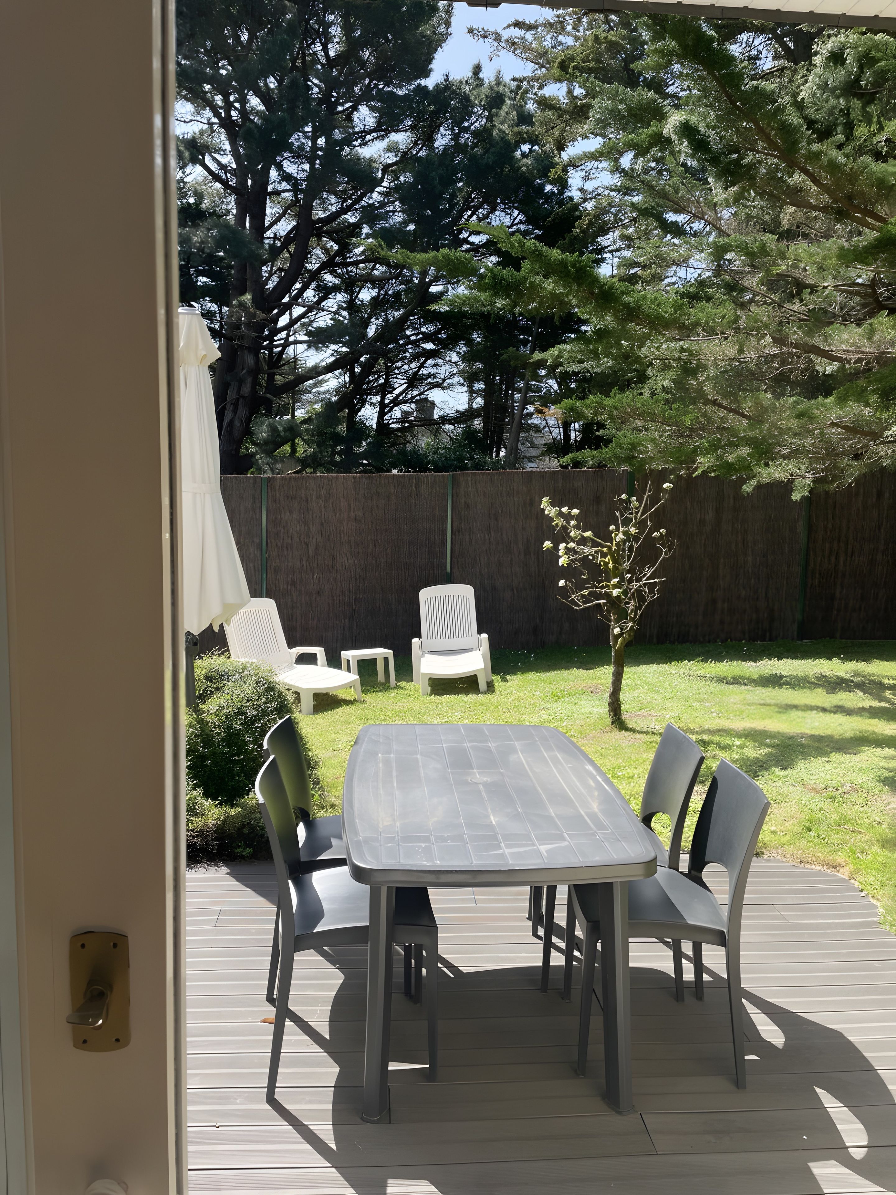 Eine Terrasse im Freien mit Tisch und Stühlen, ein Holzdeck und Blick auf einen Garten mit Zaun und Bäumen.