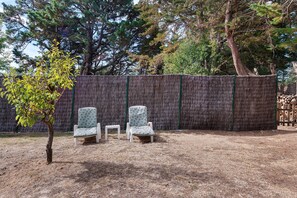 Un espace extérieur aménagé avec deux chaises et une petite table, entouré d’une clôture faite de bottes de paille et d’arbres.