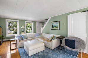A spacious living room with a sectional sofa, a small table, and a chair. There are large windows providing natural light and a view of the outdoors.